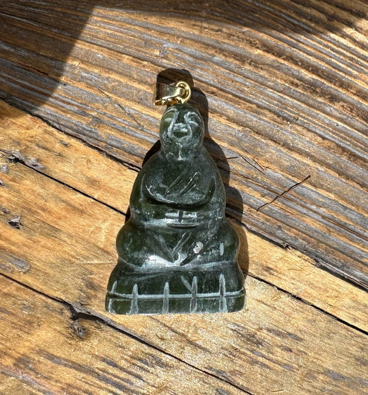 Dark jade Buddha pendant with a gold bail on a rustic wooden surface