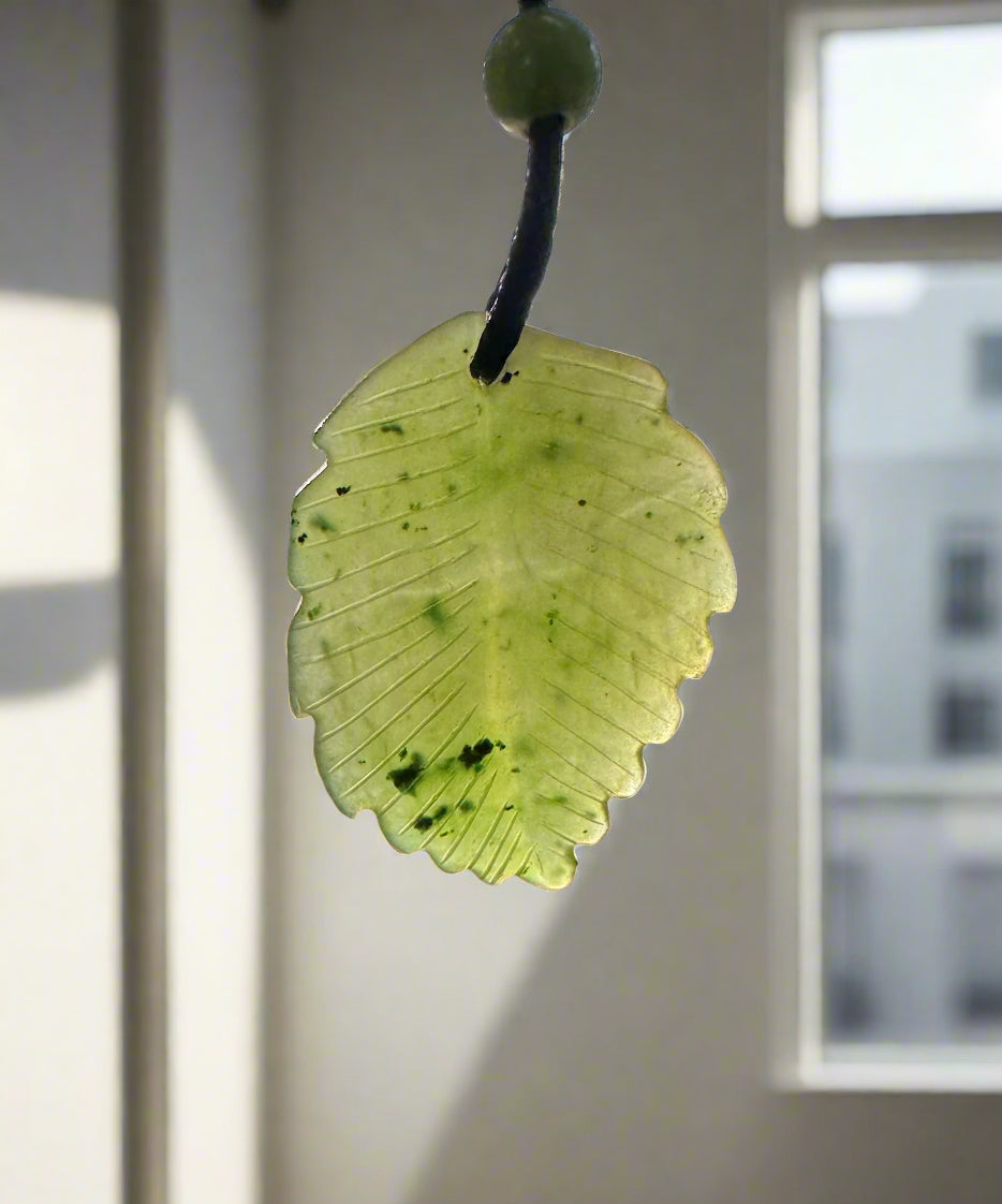 Pale green jade leaf pendant with carved veins suspended from a dark cord near a window