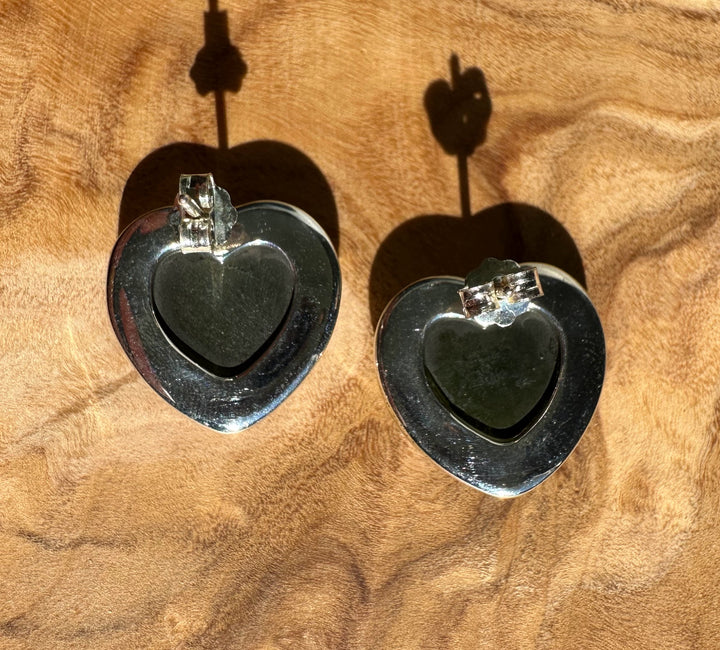 Close up of two large silver heart earrings with green center stones and posts.