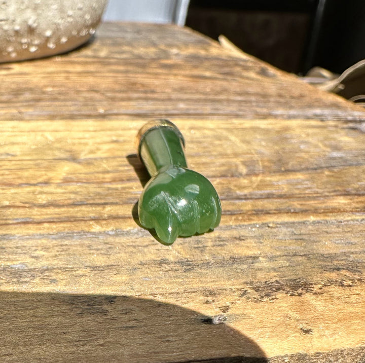 Green jade figa pendant shown from the side with a gold-tone cap and bail on a wooden surface.