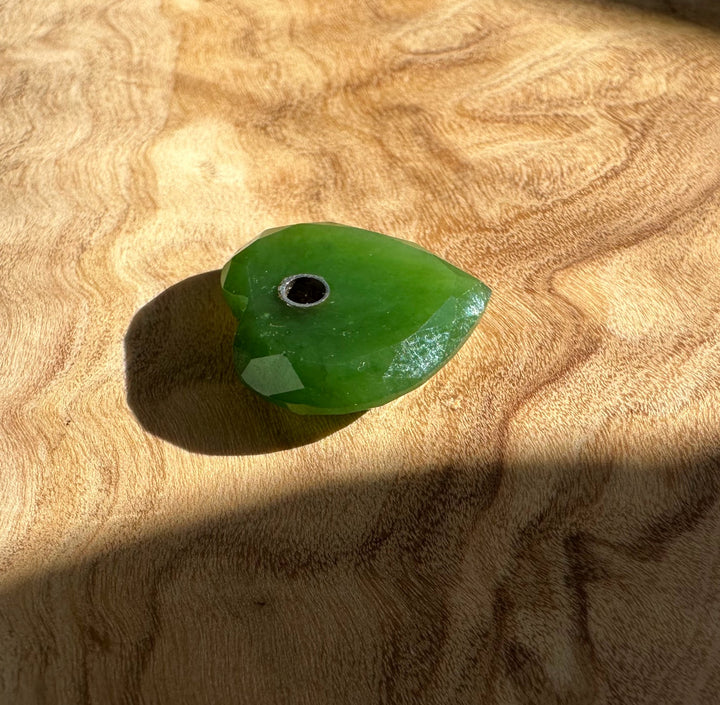 Close-up of a single faceted green jade heart bead with a drilled hole on a wood surface.