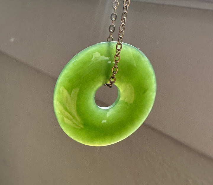 Green jade pi pendant on gold chain, with sunlight going through the jade against a grey background