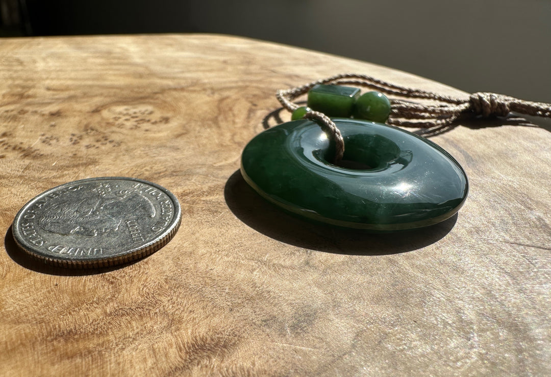 Green Jade PI Pendant on a brown cord. Resting on a wooden base, beside a US quarter for scale