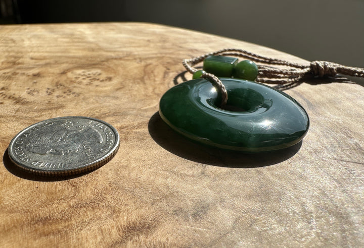 Green Jade PI Pendant on a brown cord. Resting on a wooden base, beside a US quarter for scale