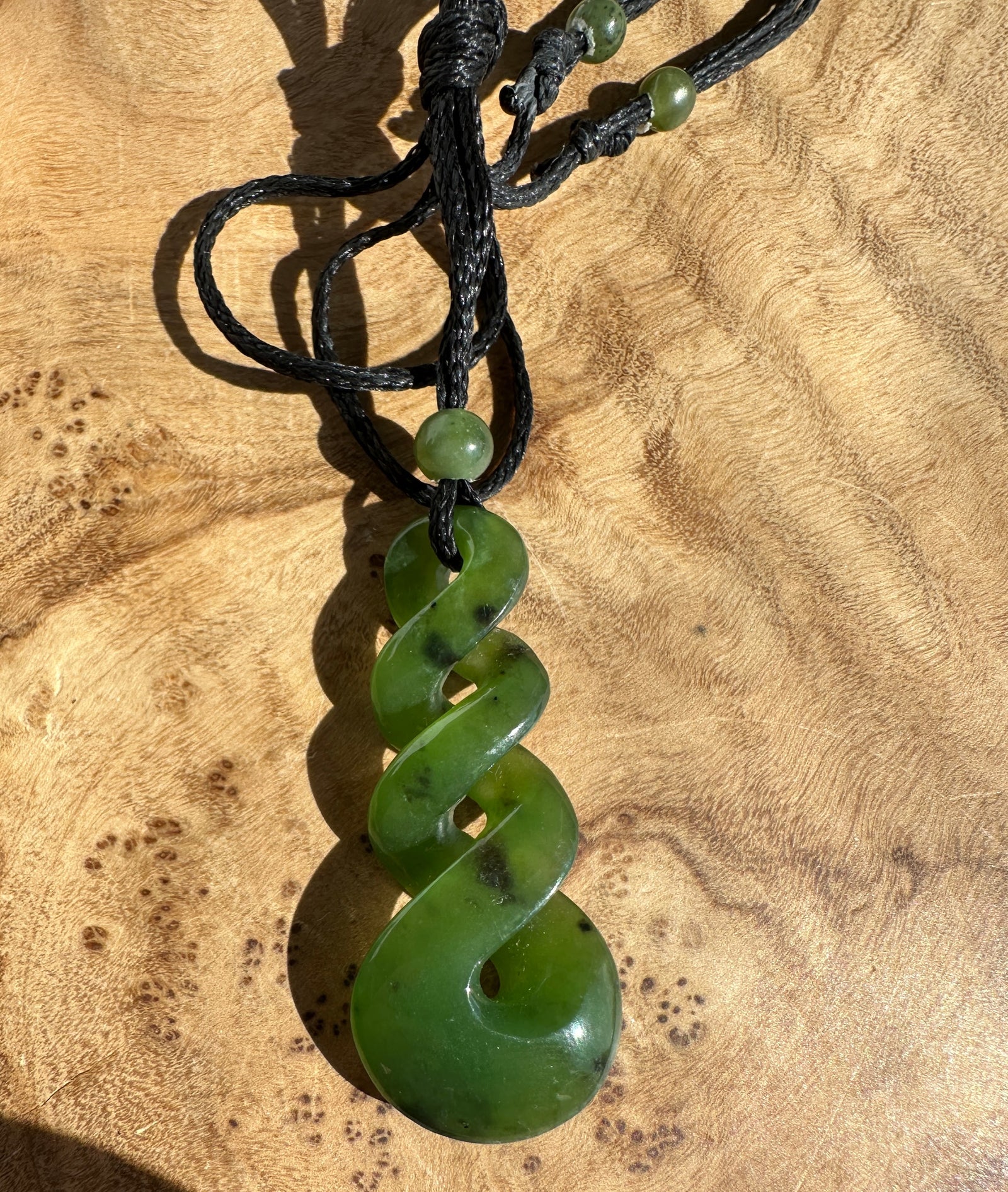 Green jade twist pendant on a black cord, laying on top of a wooden surface