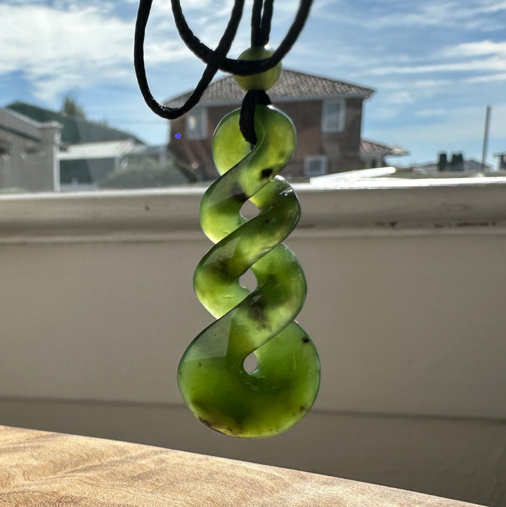 Green jade twist pendant on a black cord, infront of a sunny window 
