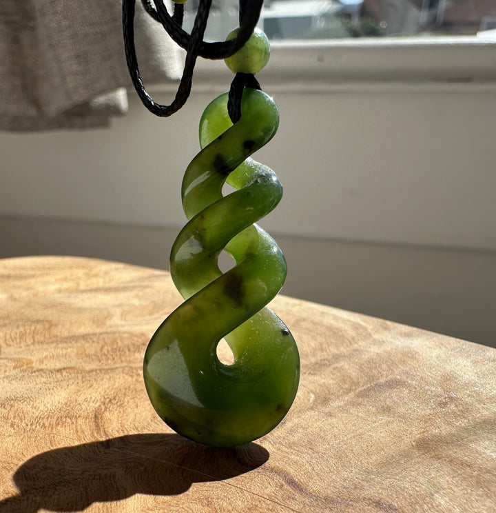 Green jade twist pendant on a black cord, laying on top of a wooden surface in front of a sunny window