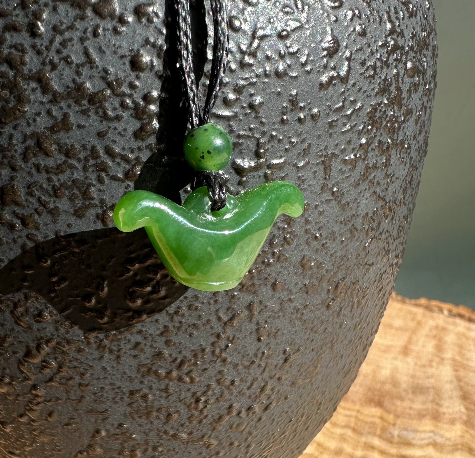 Close up of round green jade bead and curved winged pendant on a black cord against a textured stone surface