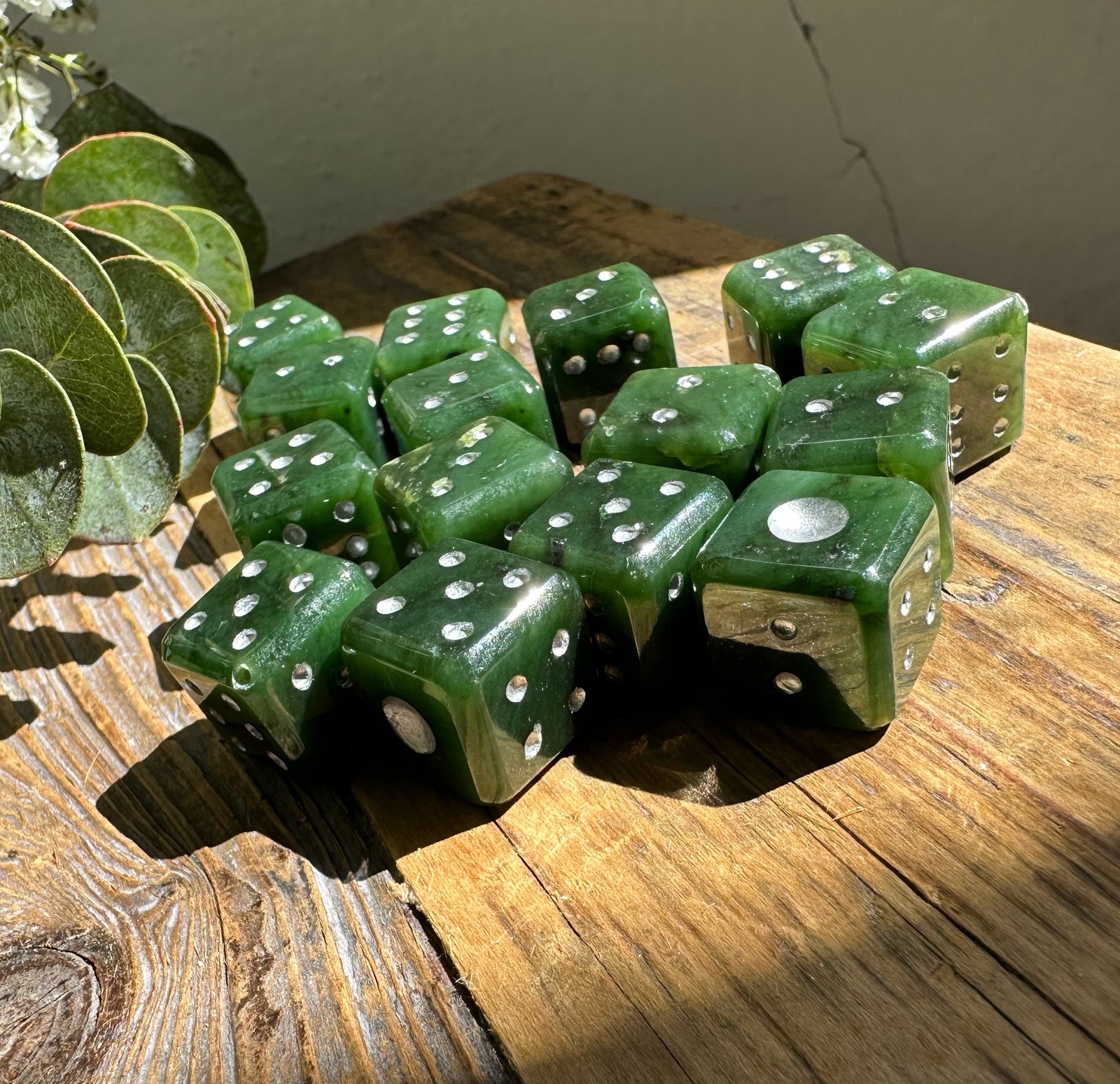 Group of green jade dice with white pips on a sunlit wooden table with greenery in the background.