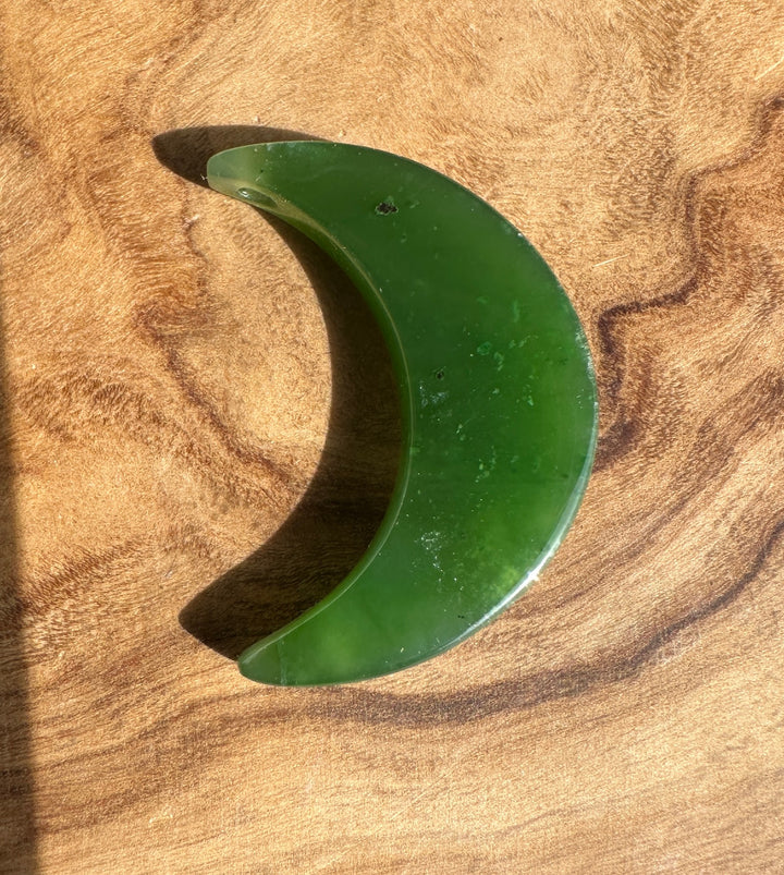 A crescent-shaped green jade pendant  polished finish, resting on a textured wooden surface.