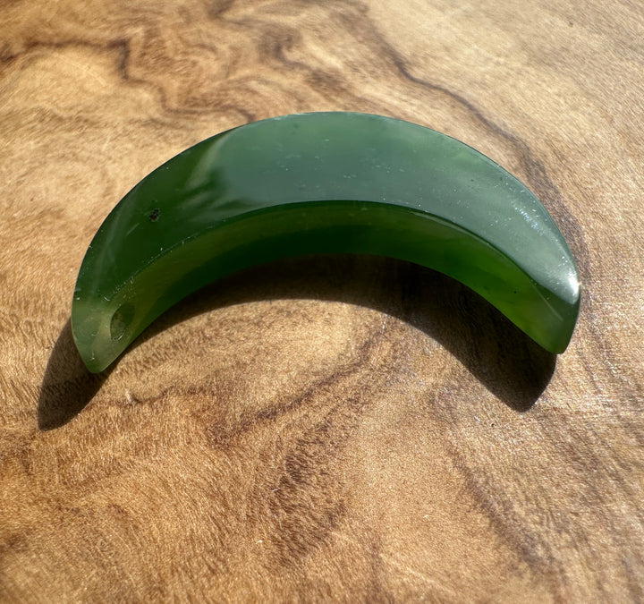 A crescent-shaped green jade accessory with a small hole on one end, resting on a textured wooden surface.