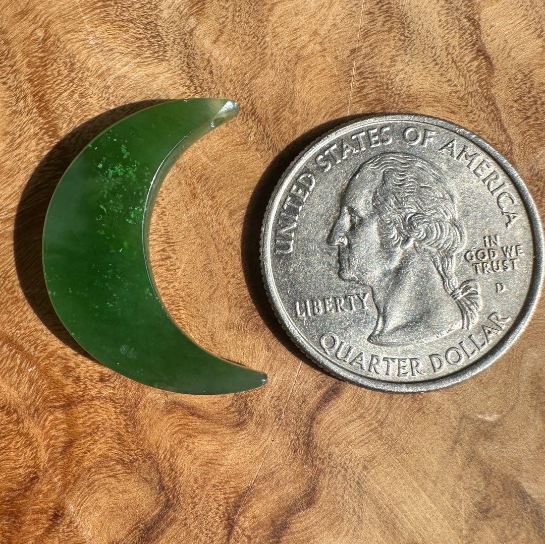 A crescent-shaped green jade piece shown next to a US quarter coin for scale, placed on a textured wooden surface.