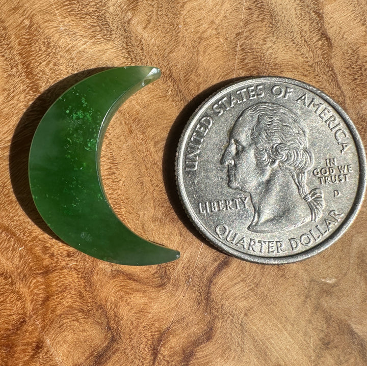 A crescent-shaped green jade piece shown next to a US quarter coin for scale, placed on a textured wooden surface.