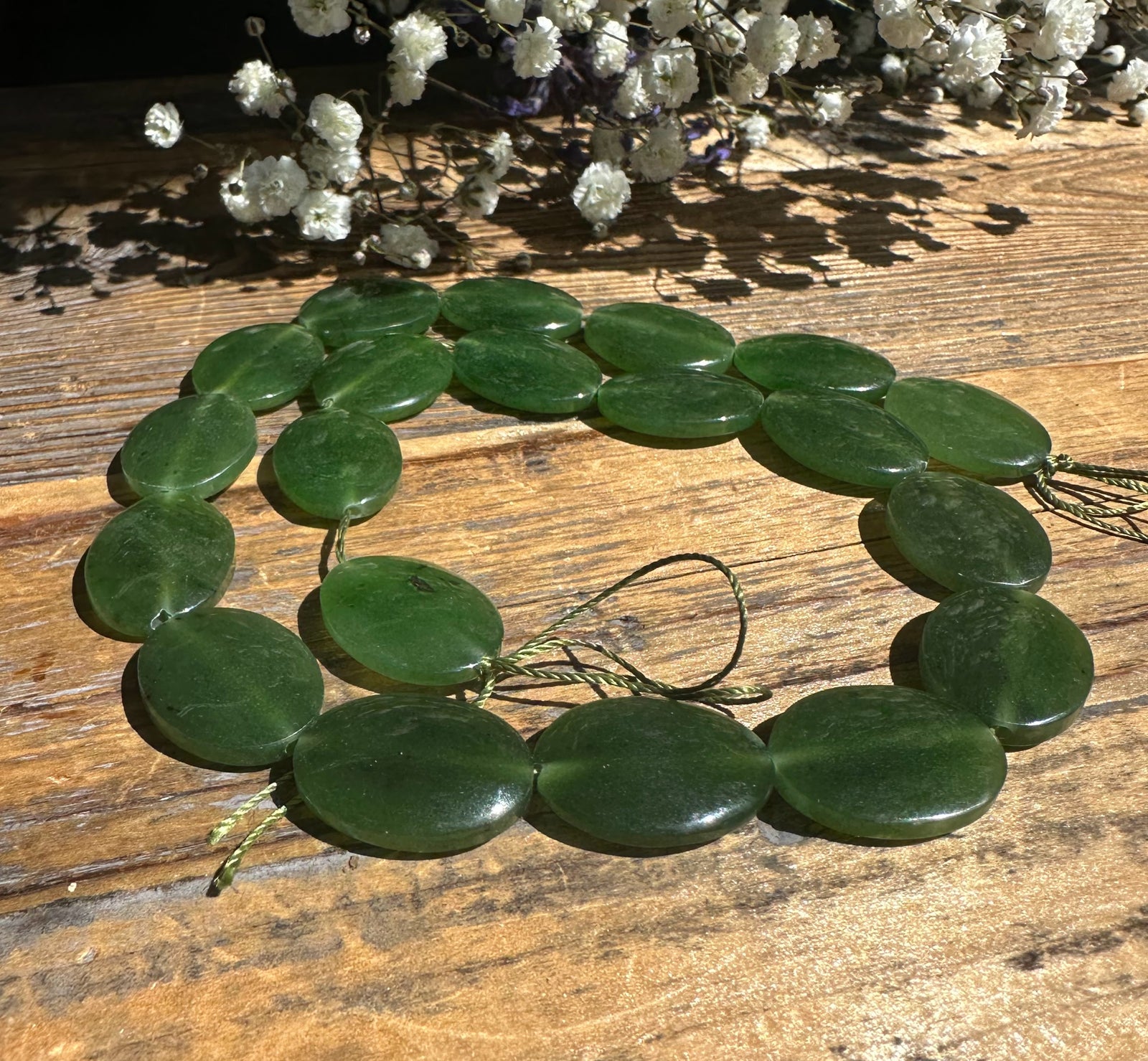 Green jade beads 20x15mm arranged as a circular necklace on a wood surface with dried white flowers in the background.