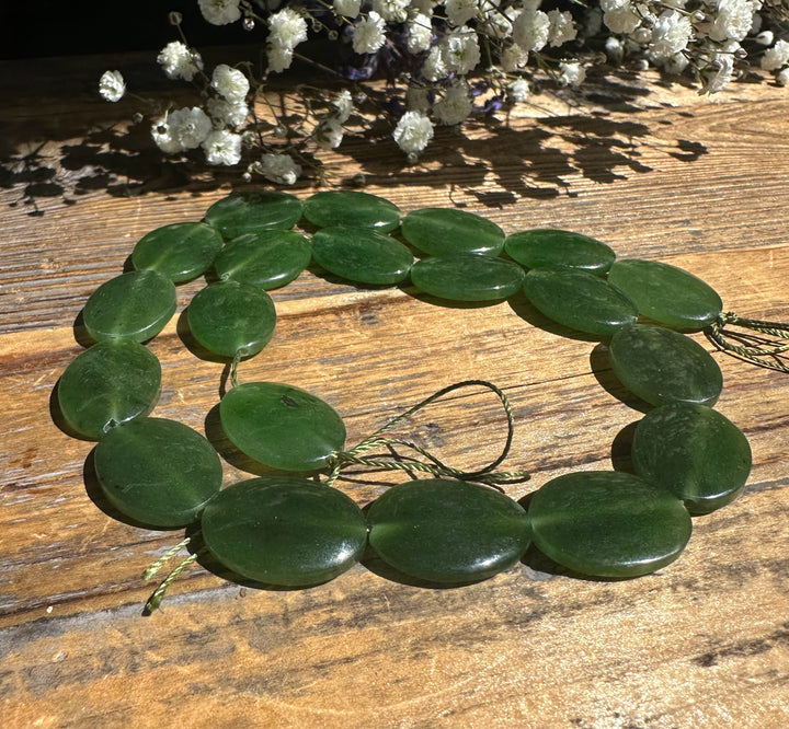 Green jade beads 20x15mm arranged as a circular necklace on a wood surface with dried white flowers in the background.