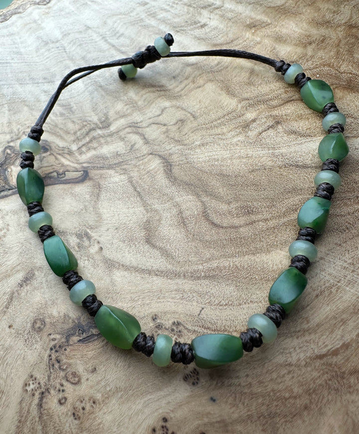 Beaded bracelet with green stones and dark braided cord resting on a wooden surface.