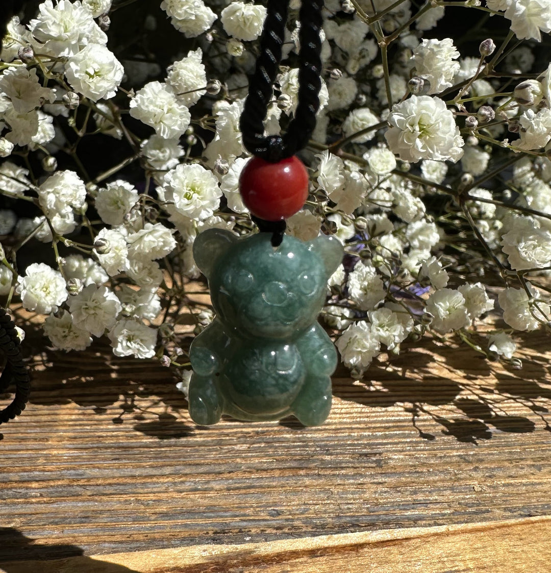 Guatemalan jadeite teddy bear pendant on a black braided cord with a red bead, set against white flowers.