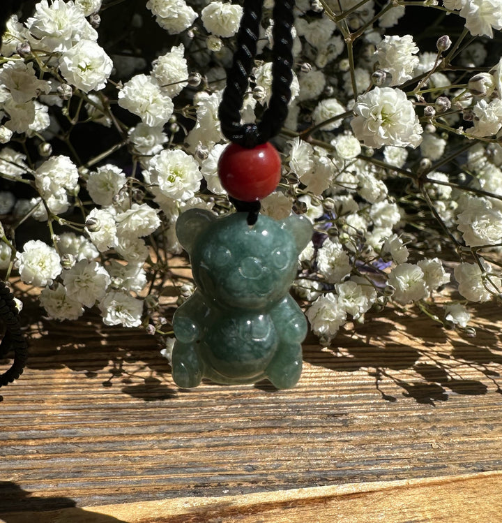 Guatemalan jadeite teddy bear pendant on a black braided cord with a red bead, set against white flowers.
