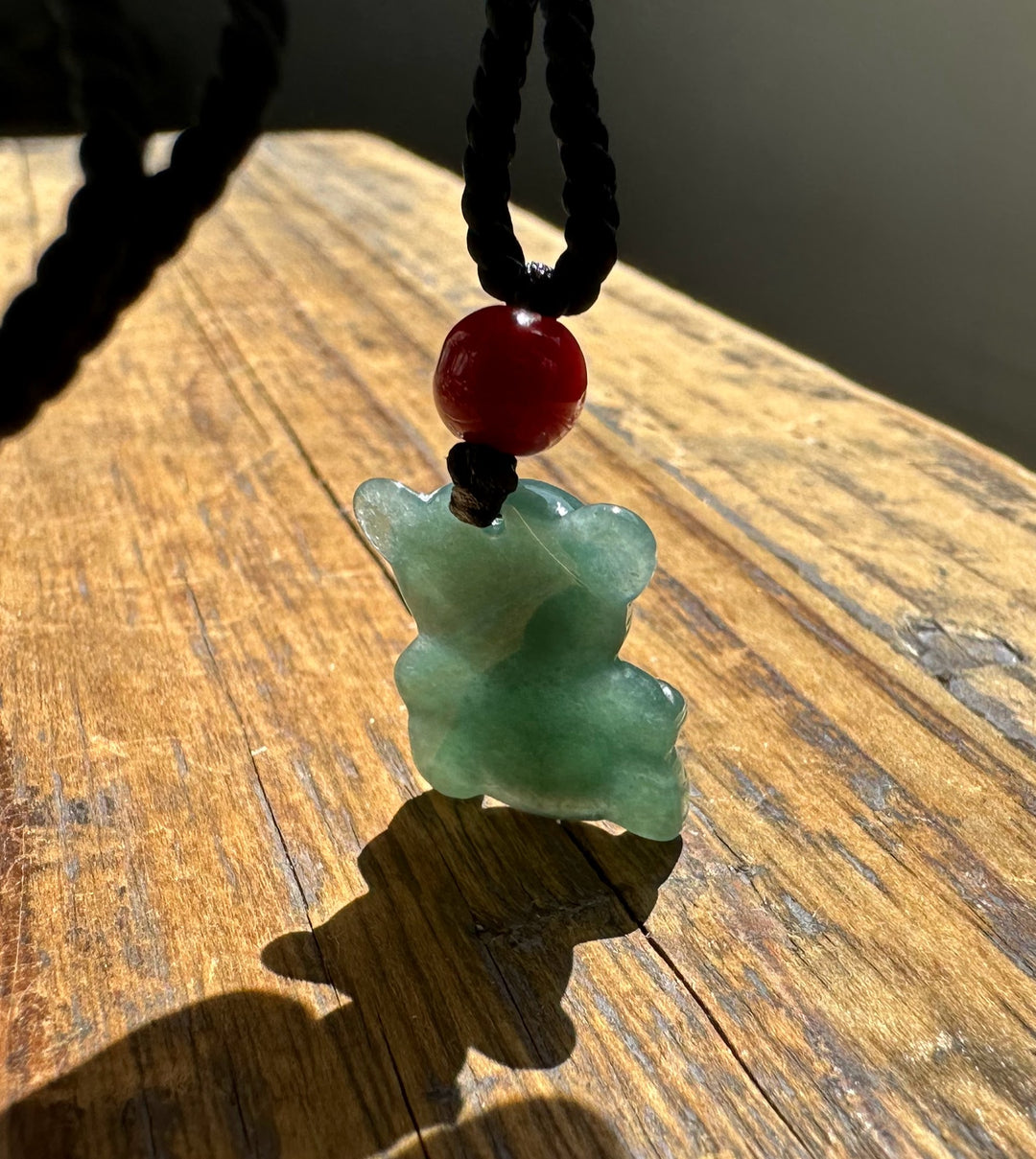 Jadeite teddy bear pendant on black cord with red bead, resting on sunlit wooden surface.