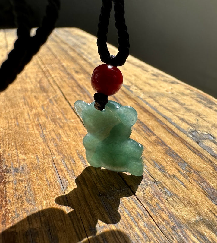 Jadeite teddy bear pendant on black cord with red bead, resting on sunlit wooden surface.