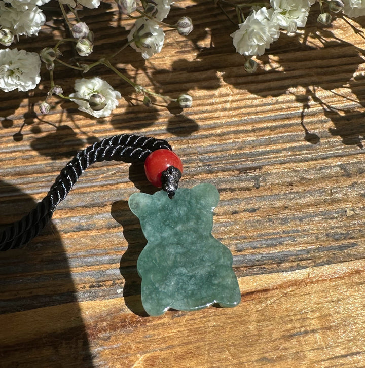 Green jadeite teddy bear pendant on wooden surface with white flowers around and braided cord nearby.