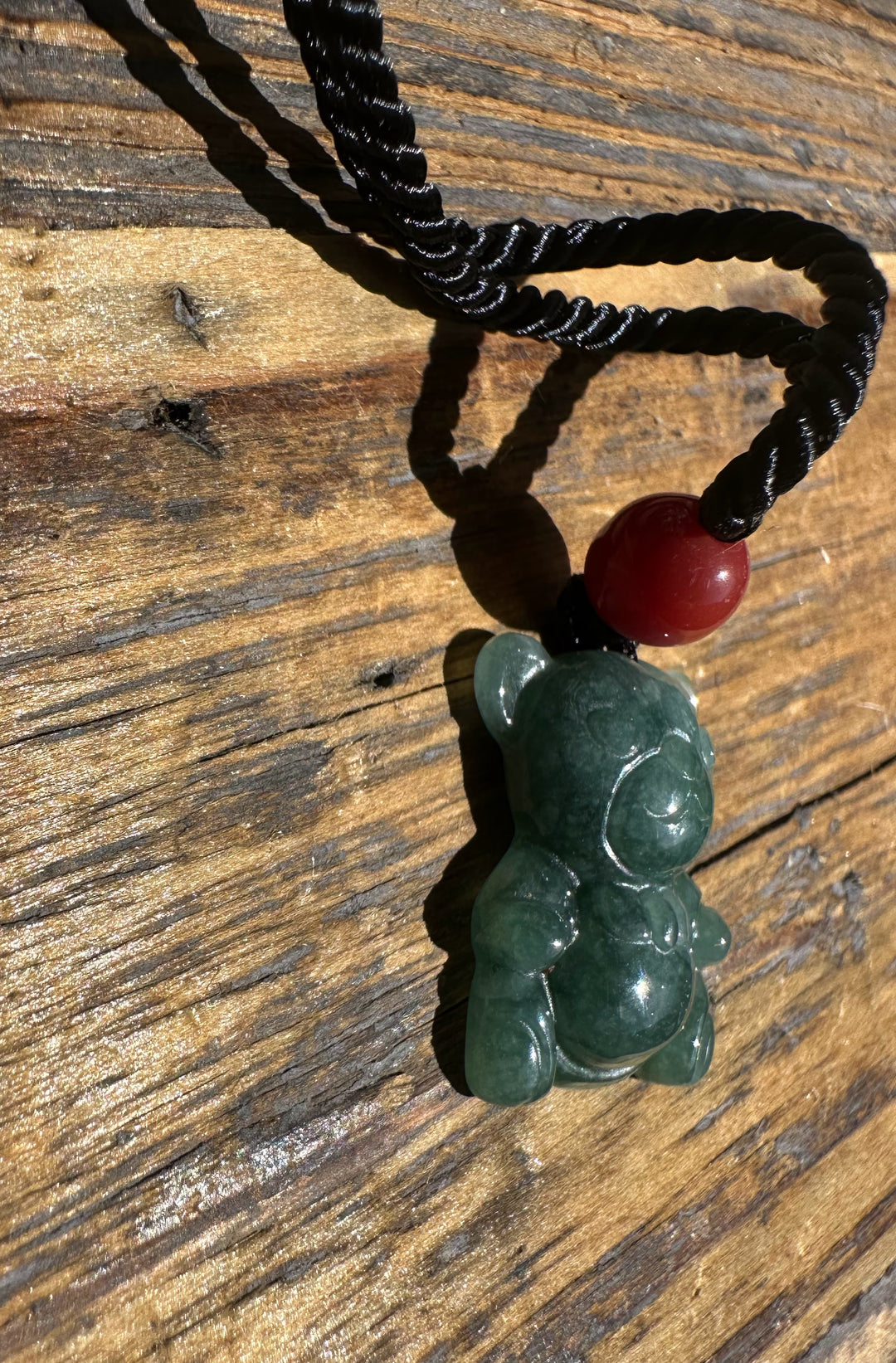 Guatemalan jadeite teddy bear pendant with a red bead on a black braided cord placed on wood