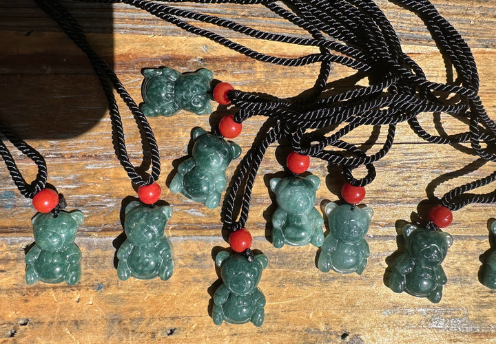 Multiple Guatemalan jadeite teddy bear pendants on black cords with red beads on a wooden surface