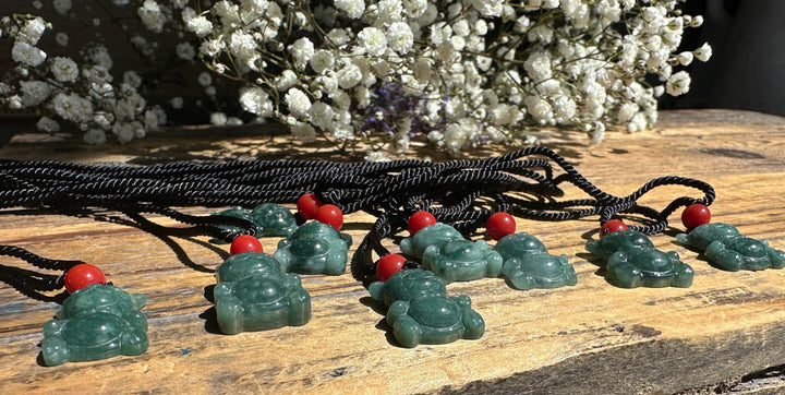 Row of Guatemalan jadeite teddy bear pendants on black cords with red beads on a wooden table with white flowers in background