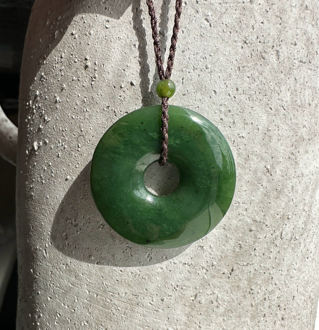 Dark green jade donut pendant suspended on brown braided cord against a textured stone background