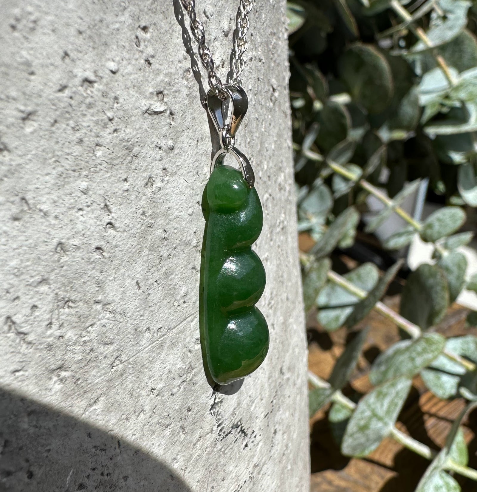 Close up of a jade pea pod pendant hanging from a silver chain against a rough stone surface.