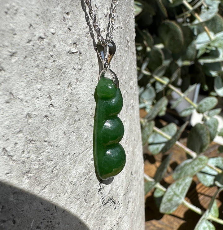 Close up of a jade pea pod pendant hanging from a silver chain against a rough stone surface.