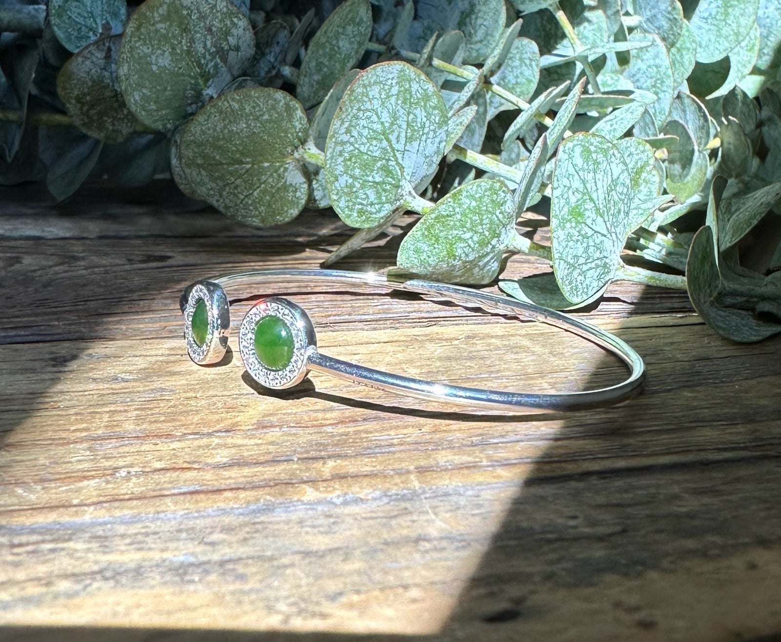 Sterling silver bracelet with two green cabochon stones in round diamond halos on a rustic wooden surface.