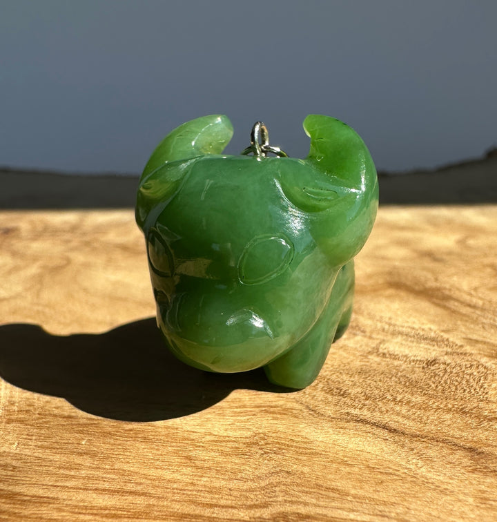 A carved green jade animal pendant, shaped like a buffalo or ox, with a small silver loop at the top, resting on a wooden surface in natural light.