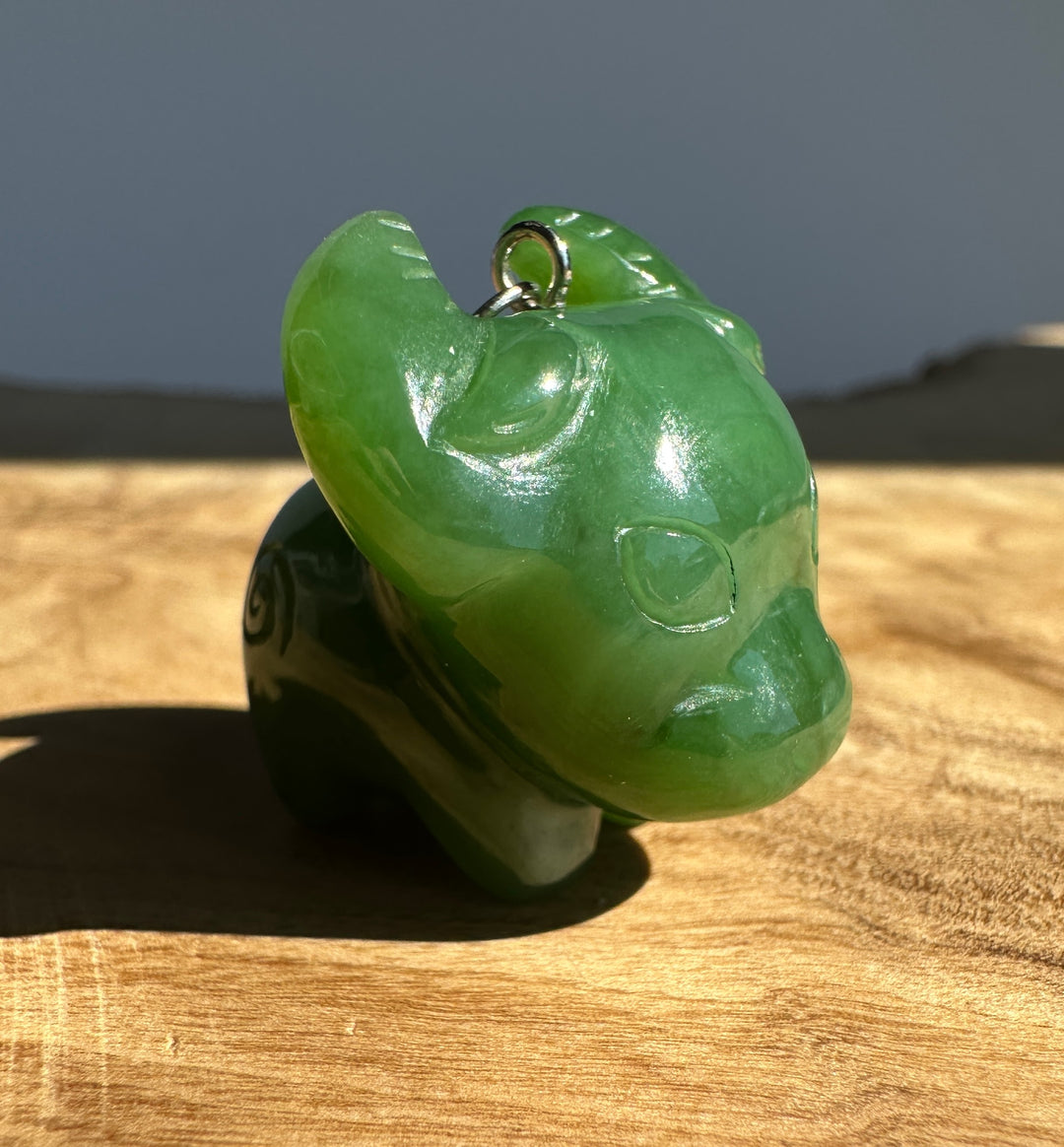 A carved green jade animal pendant, shaped like a buffalo or ox, with a small silver loop at the top, resting on a wooden surface in natural light.