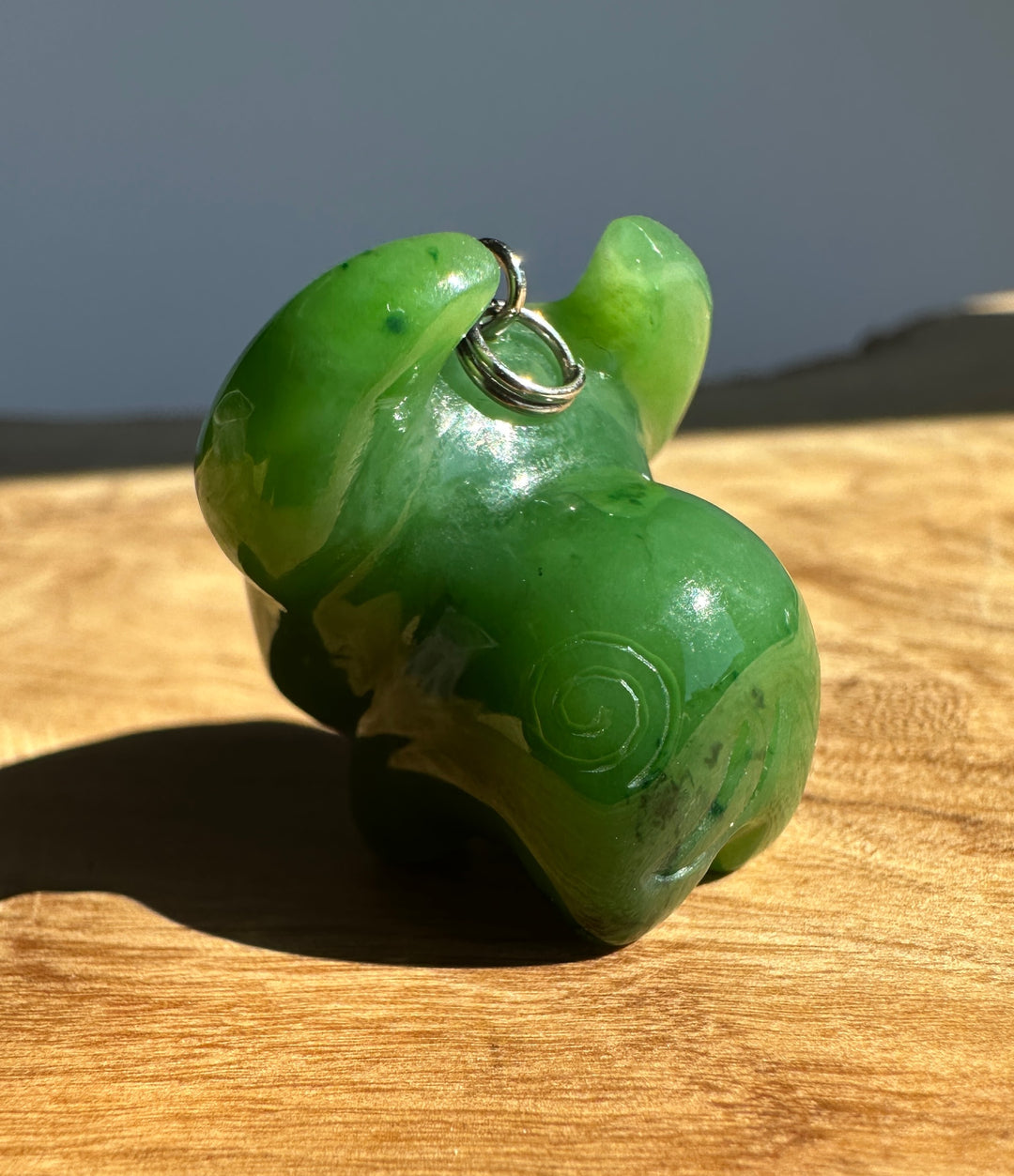 A carved green jade animal pendant, shaped like a buffalo or ox, with a small silver loop at the top, resting on a wooden surface in natural light.