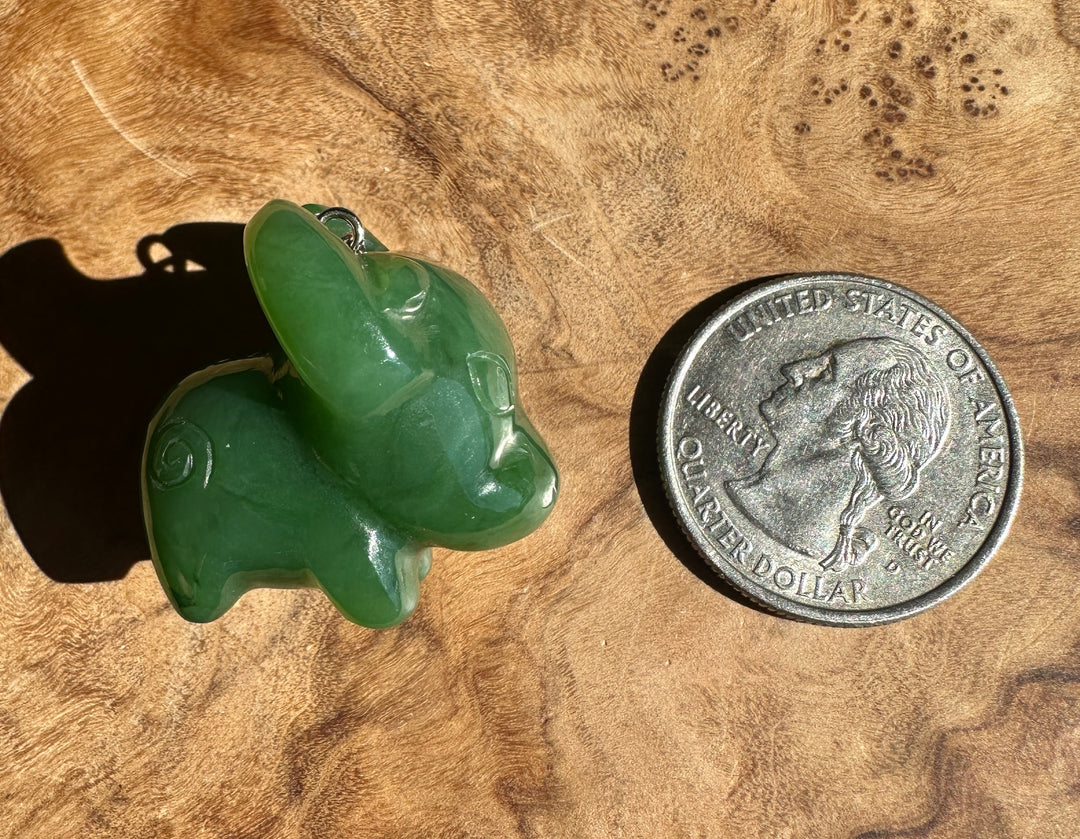 A carved green jade animal pendant, shaped like a buffalo or ox, with a small silver loop at the top, resting on a wooden surface in natural light next to a US quarter