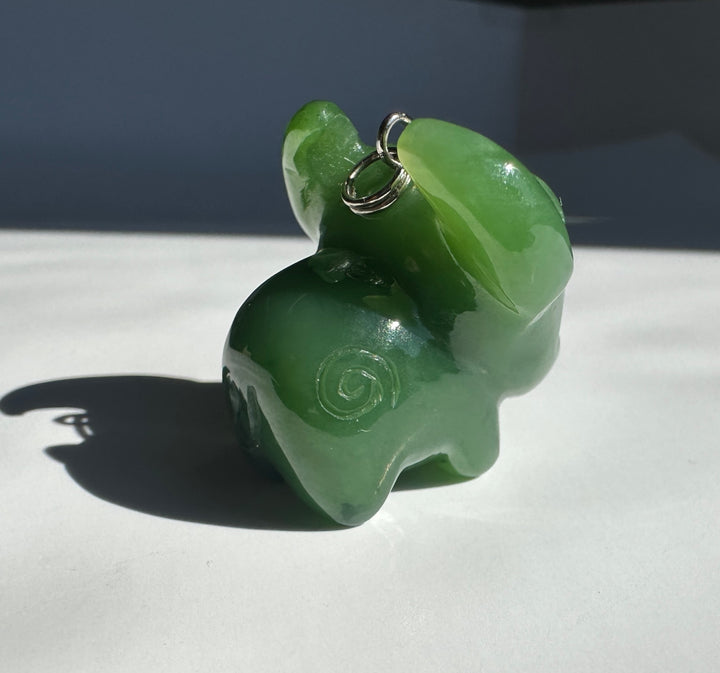A small carved green stone animal pendant or charm with a silver metal loop at the top, photographed on a white surface with strong sunlight and shadow.