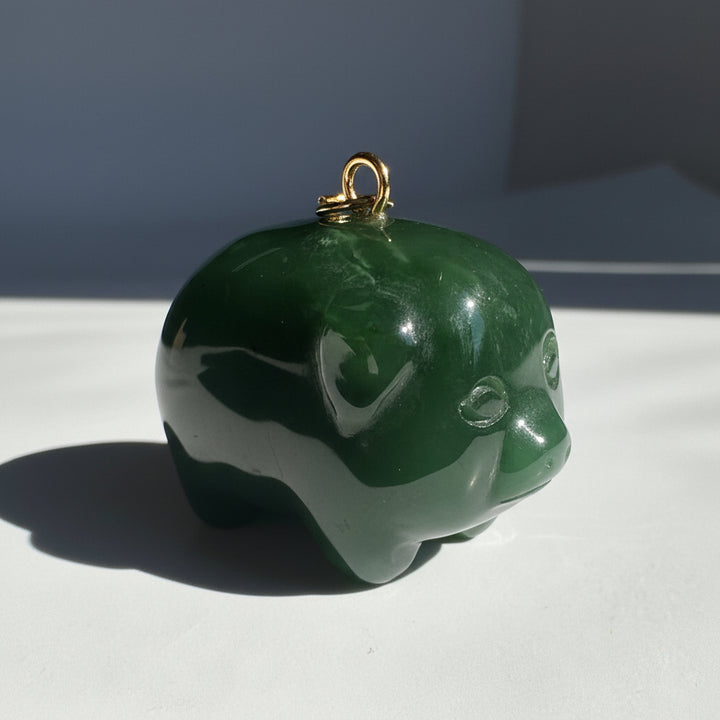 A polished dark green pig-shaped pendant or charm with a small gold loop at the top, photographed on a white surface in bright sunlight.
