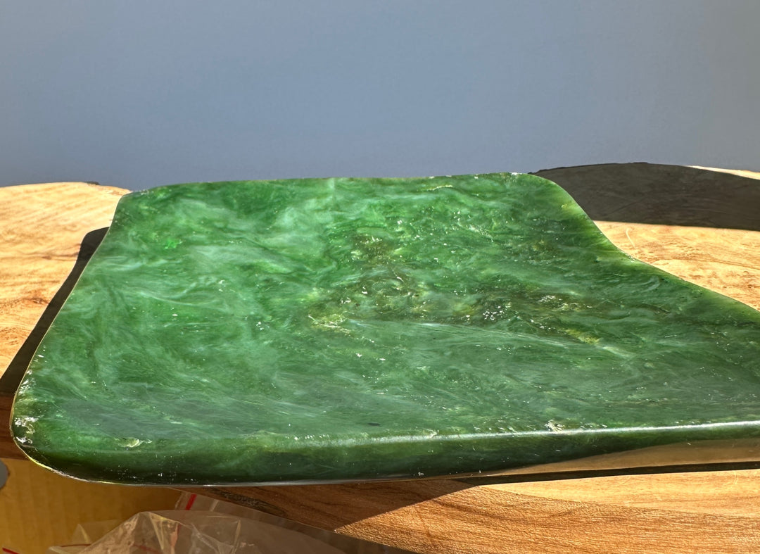 A polished green jade slab with natural marbled patterns, resting on a wooden surface against a blue-gray background.