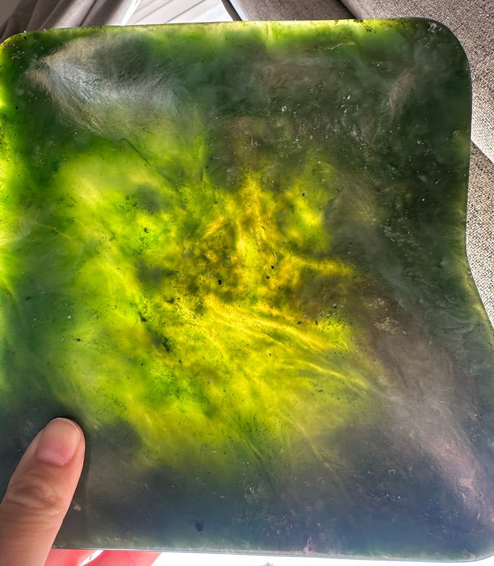 A hand holding a square translucent green stone slab with a marbled pattern of bright yellow-green and dark green, photographed indoors.