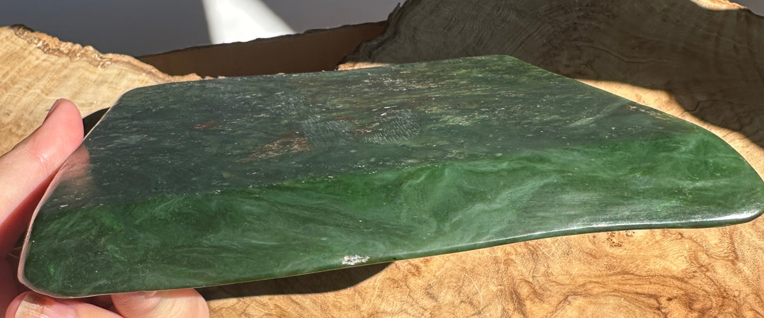 A polished green stone jade rectangular slab with marbled natural patterns and small orange-brown inclusions, held in a person&