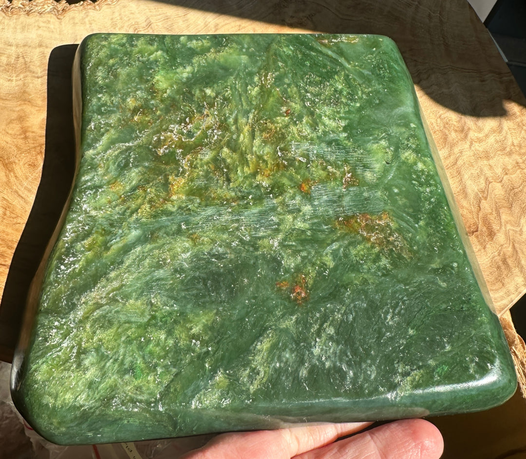 A polished green stone jade rectangular slab with marbled natural patterns and small orange-brown inclusions, held in a person&