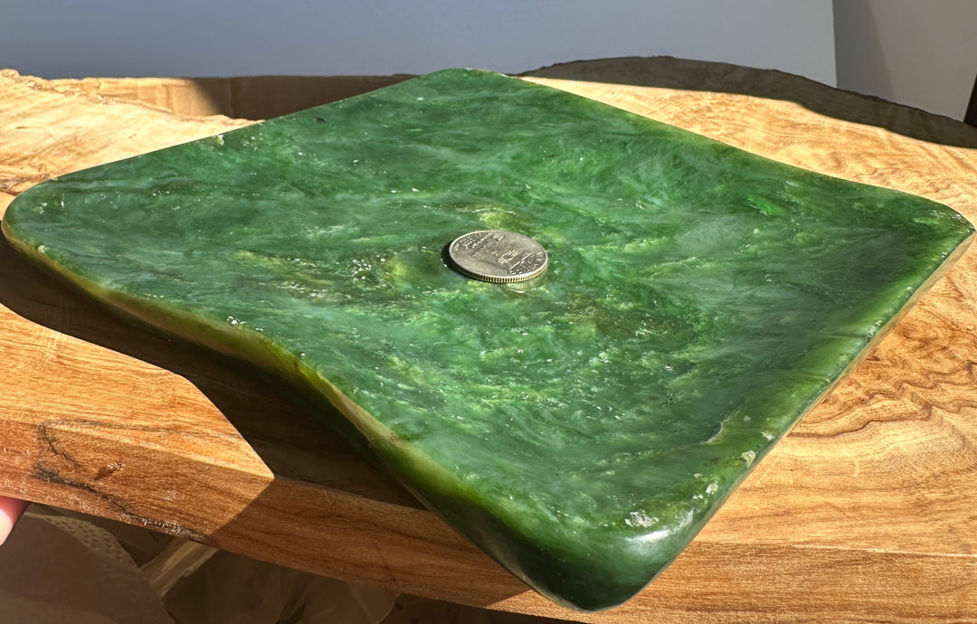 A square green stone or polished wood serving board with rounded corners and a small coin placed on top for scale, resting on a wooden surface.