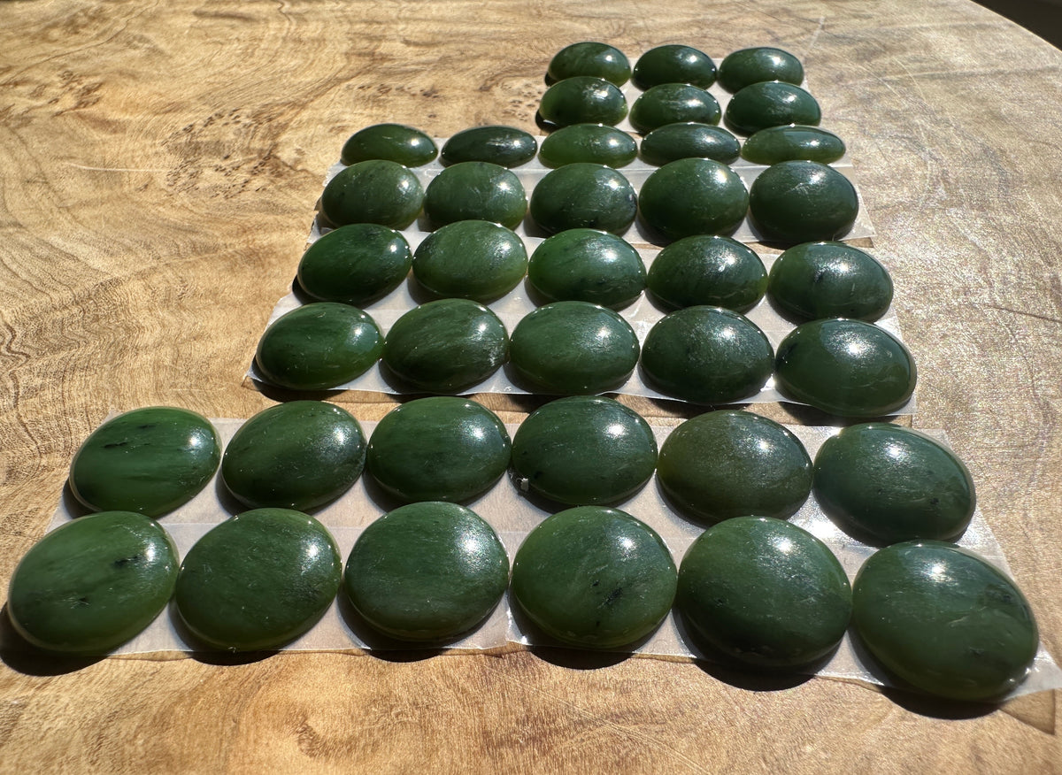 A set of polished dark green jade cabochons stones arranged in rows on a light wood background.