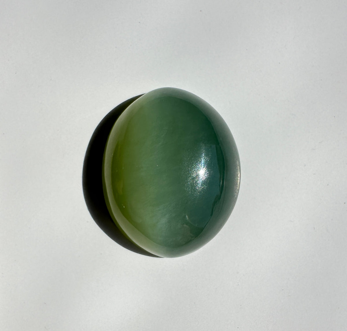A polished oval green stone or cabochon with a glossy finish, casting a soft shadow on a plain light background.