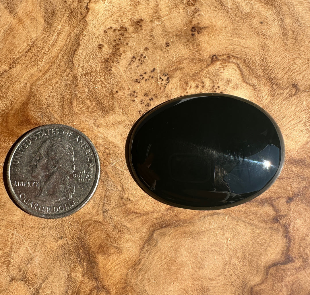A polished black oval cabochon gemstone shown next to a US quarter for scale, placed on a textured burl wood surface.