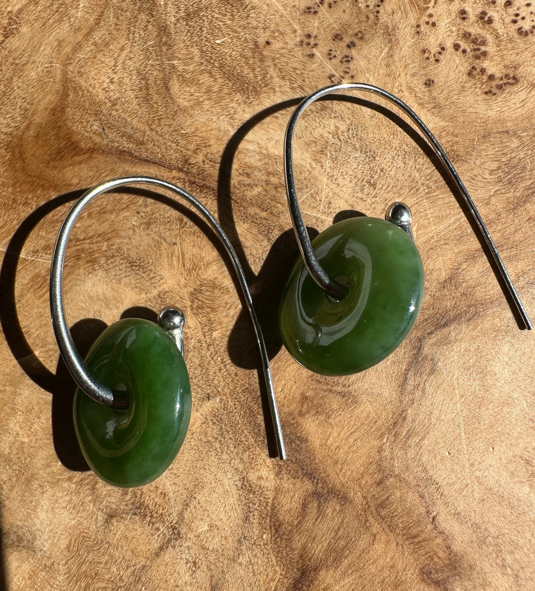 A pair of handmade earrings featuring polished green stone discs with a central hole, suspended from thin silver-colored metal wires, displayed on a textured wooden surface.