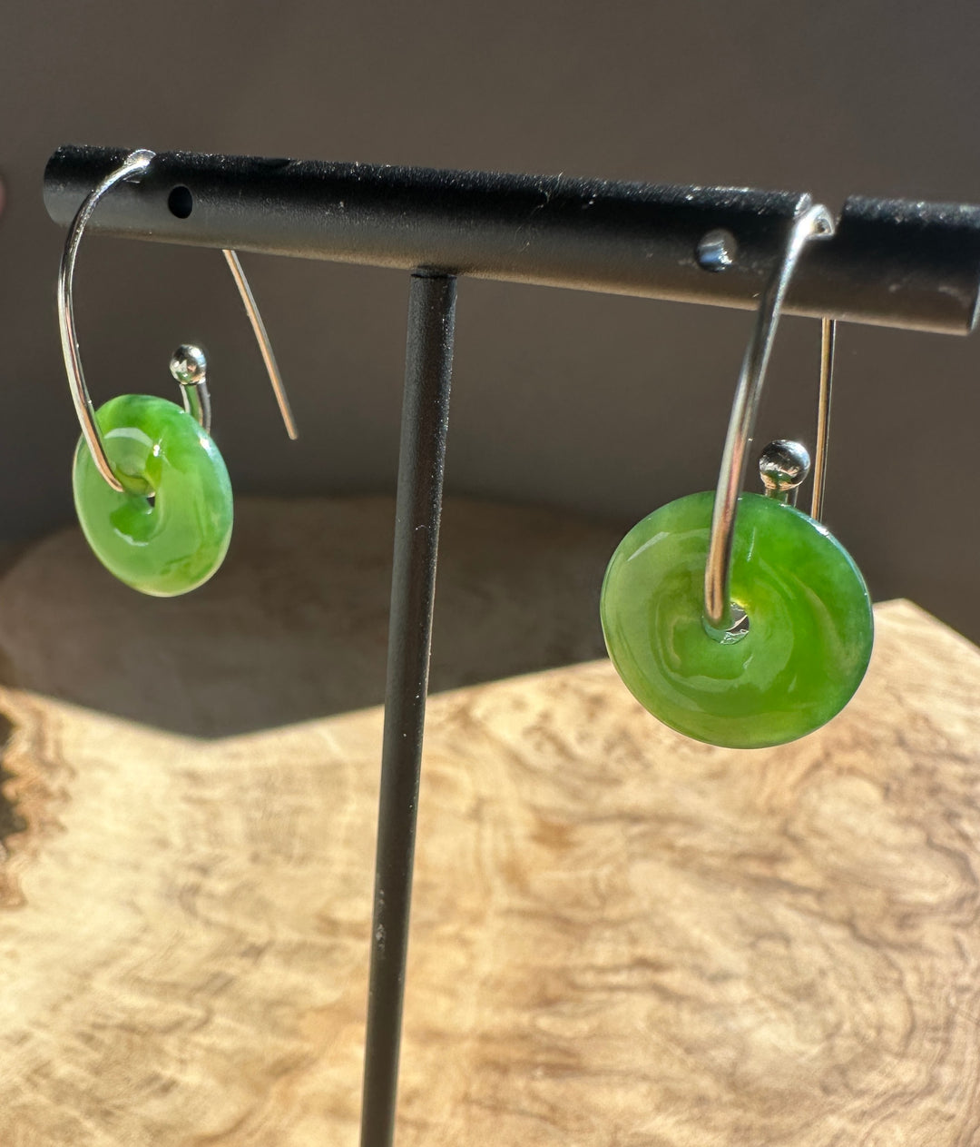 A pair of green disc-shaped earrings with silver hooks, displayed on a black jewelry stand over a wood slice background.
