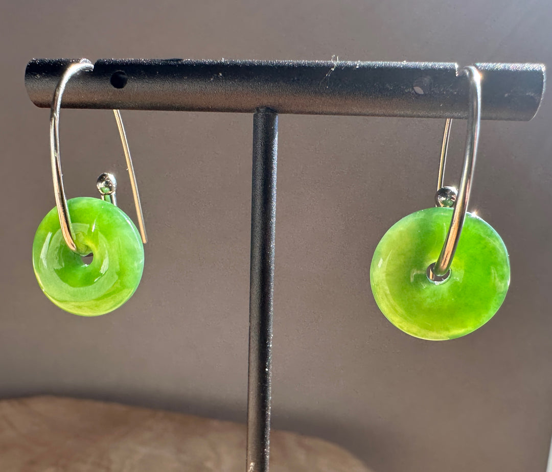A pair of green disc-shaped earrings with silver-toned hooks, displayed on a black jewelry stand against a neutral gray background.