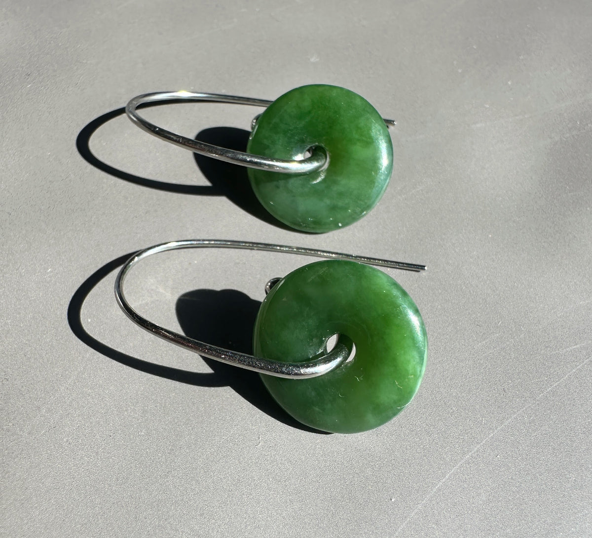 A pair of green stone donut earrings with silver wire hooks, displayed on a neutral gray surface with sharp shadows.